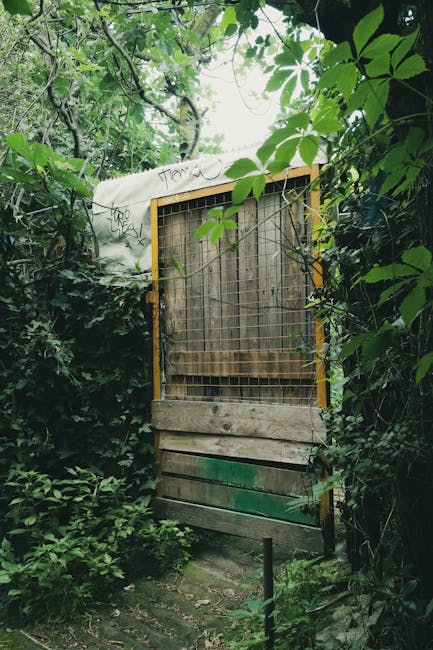 A small metal utility box covered in multicolored graffiti tags including orange, blue, pink, and black, is situated amidst dense green foliage in a garden setting. Surrounding the box are variegated