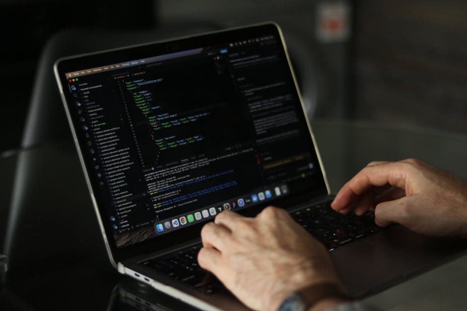 A person working outdoors on a laptop, with the screen displaying lines of code in a dark-themed programming interface. The individual is using their right hand to operate the trackpad and their left 